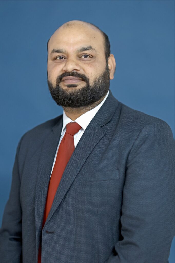 Portrait of Lalit Puri, Co-Founder of Indian Adventure Fusion, wearing a dark suit with a red tie against a blue background.