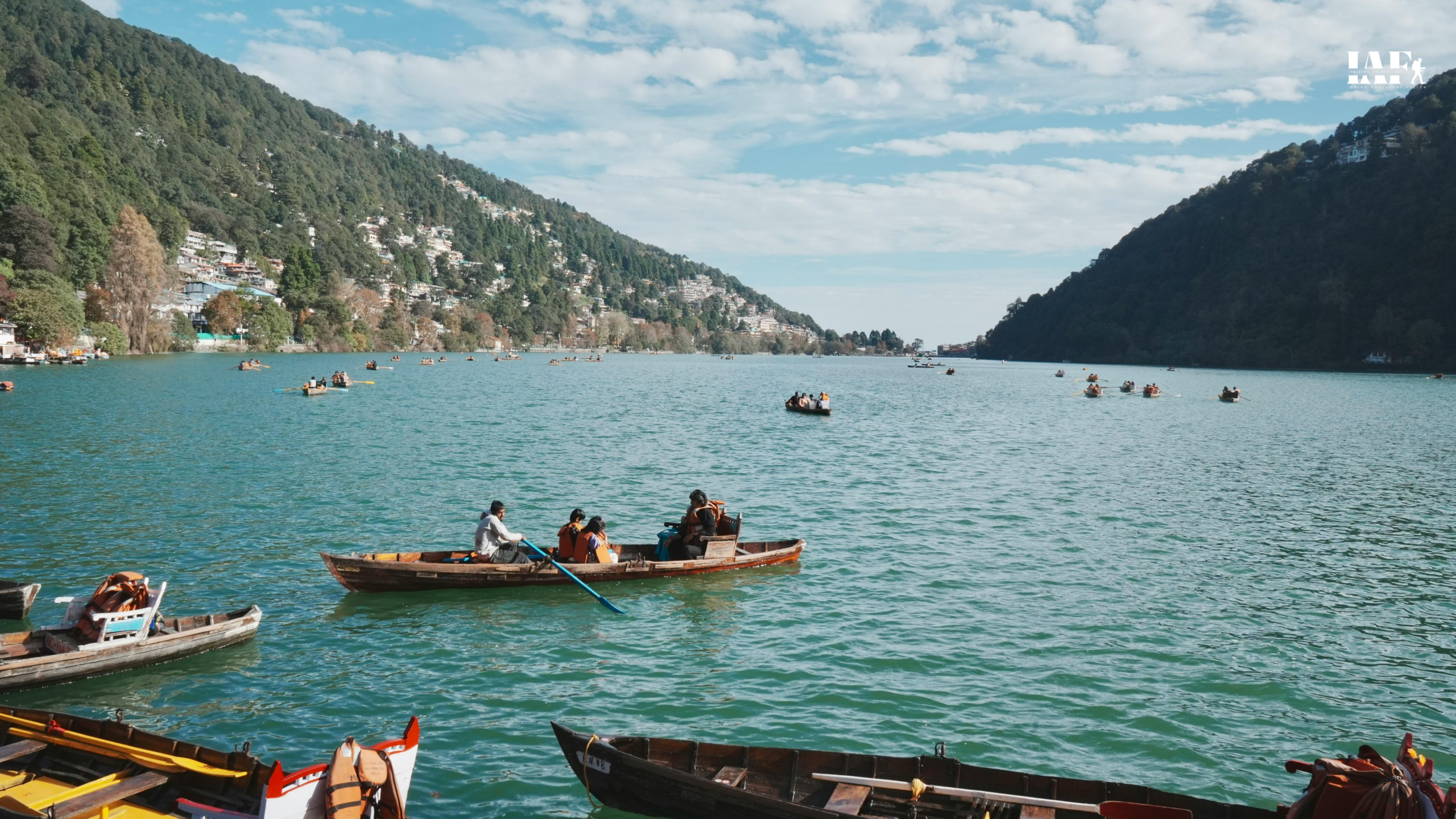 Tourists enjoying boat rides on Naini Lake in Nainital, Uttarakhand, surrounded by lush green hills and hillside houses under a partly cloudy sky.